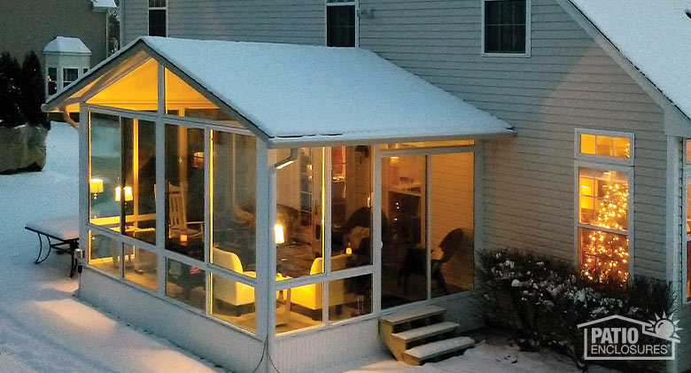 A gable-roofed sunroom at dusk with snow on the roof and on the ground.