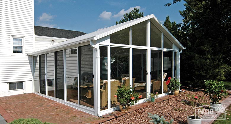 A white sunroom with gable roof and glass wings, patio doors all around. Garden with roses in front.