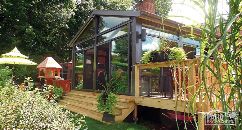A brown sunroom with gable roof, glass wings, and transom. Wooden steps in front lead down to the yard and gardens.