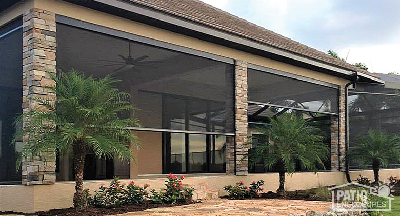 A covered patio with stone columns and dark retractable screens partly down. Rose bushes and palm trees in front.