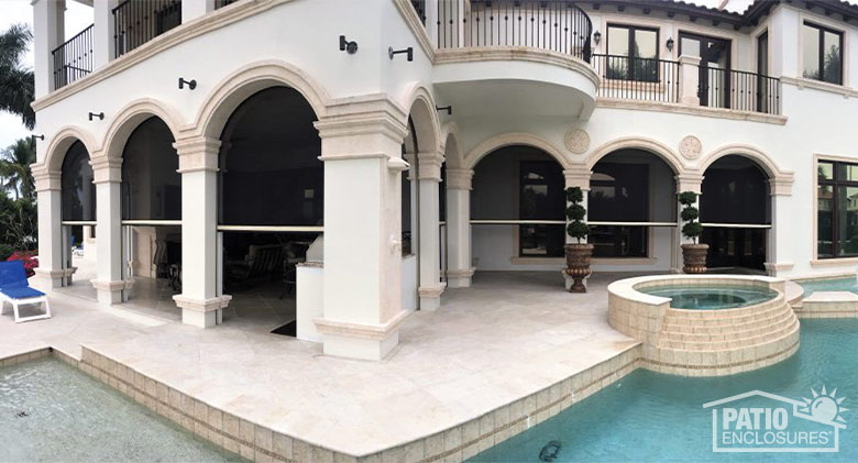 Arched openings in a covered patio are covered halfway by black retractable screens. A pool in the foreground.