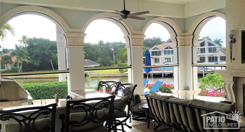 A covered patio with dining and seating areas and arched openings halfway covered with black motorized screens.
