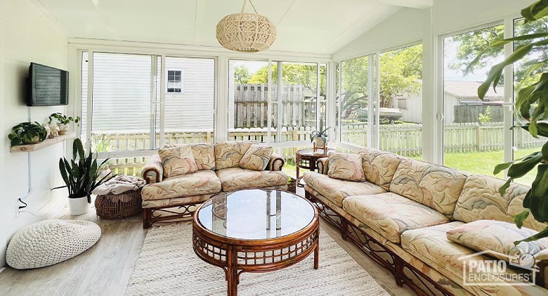 Interior of a large glass sunroom with comfortable seating, round coffee table, and wall-mounted television.