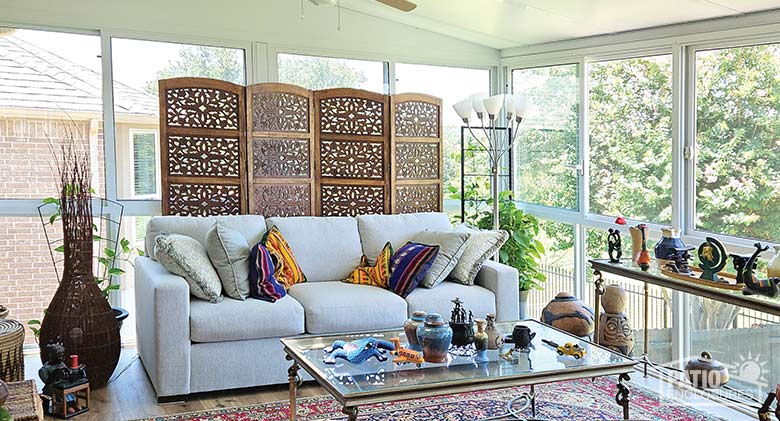 Inside of a glass sunroom: toys on a glass coffee table, gray couch with bright pillows in front of a carved room screen.