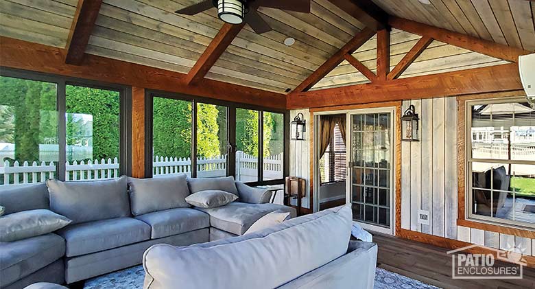 The rustic interior of a porch enclosure with wood-paneled walls, wood trim, ceiling fan, and large, gray, overstuffed sofas.