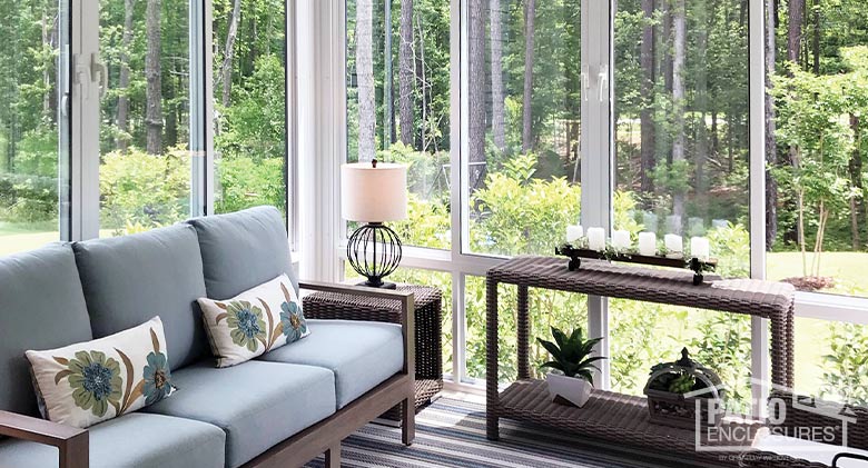 comfortable, modern interior of a white sunroom with gray couch, lamp, brown wicker side tables, view of backyard and woods