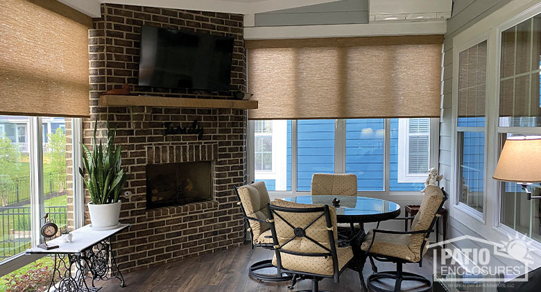 interior of glass porch enclosure, fireplace in corner with TV mounted above, dining table and shades partially drawn