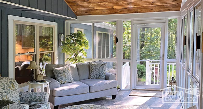 Interior of a sunroom with blue upholstered couch; print chair, ottoman and pillows; tiled floor; and sun streaming in.