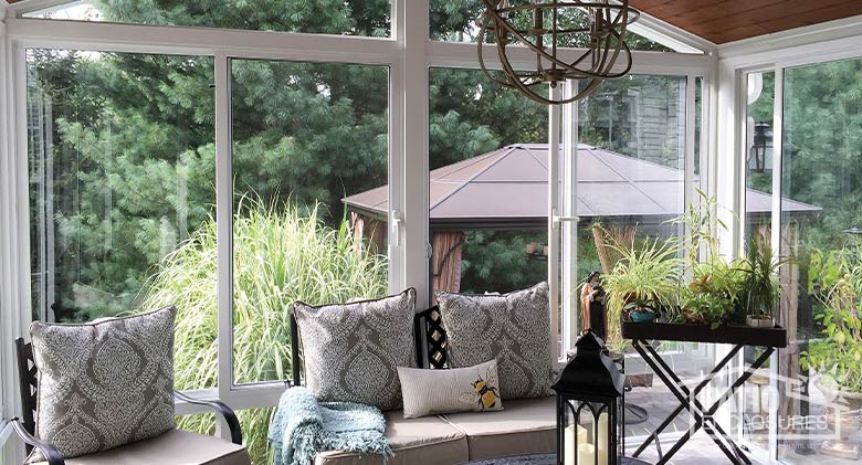 Interior of a white sunroom with comfortable furniture, pillows and plants and a view of the landscaping and gazebo outside. 