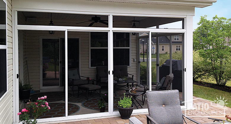 Exterior of a screened-in corner patio with white frame, transoms and a paver patio with furniture and plants outside.