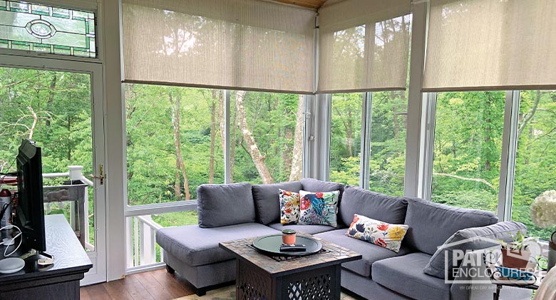 Interior of a sunroom overlooking a wooded area. An L-shaped gray couch is in the corner with a coffee table in front.