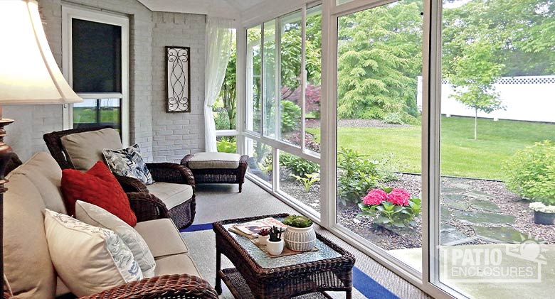 Overstuffed wicker couch and chair with pillows face glass sunroom windows overlooking a well-tended flower bed and yard.