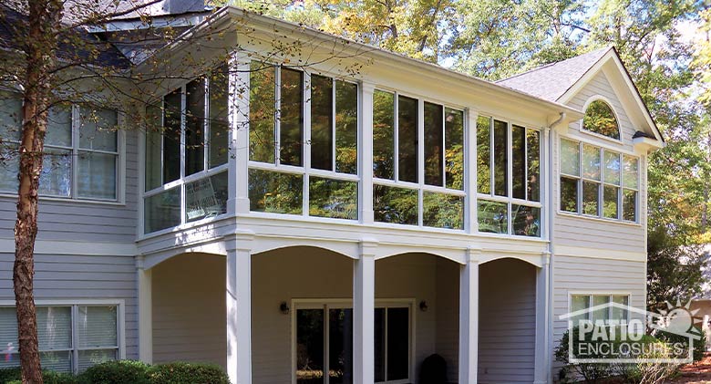 A second-story white sunroom with open patio beneath it on a gray two-story home surrounded by large trees.