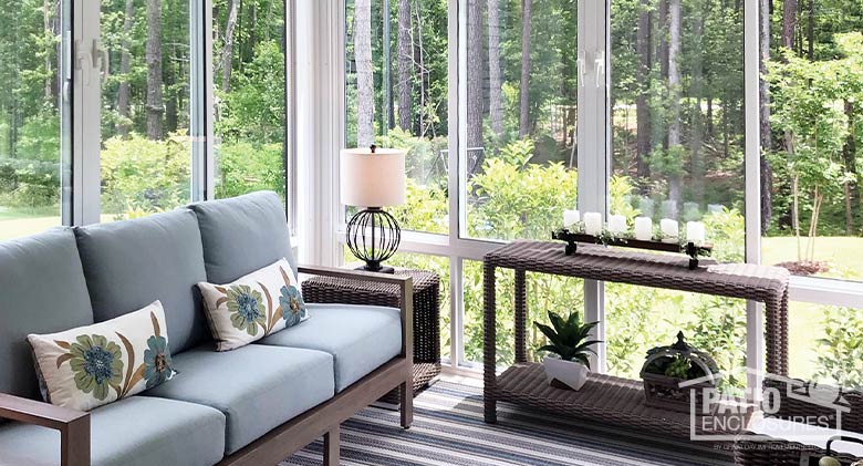 A blue couch with floral pillows and wicker side tables inside a white sunroom overlooking a backyard and woods.