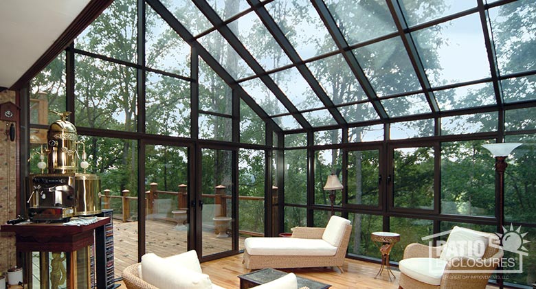 Beige wicker furniture with white cushions inside a soaring brown solarium with glass roof and walls and wood floor.