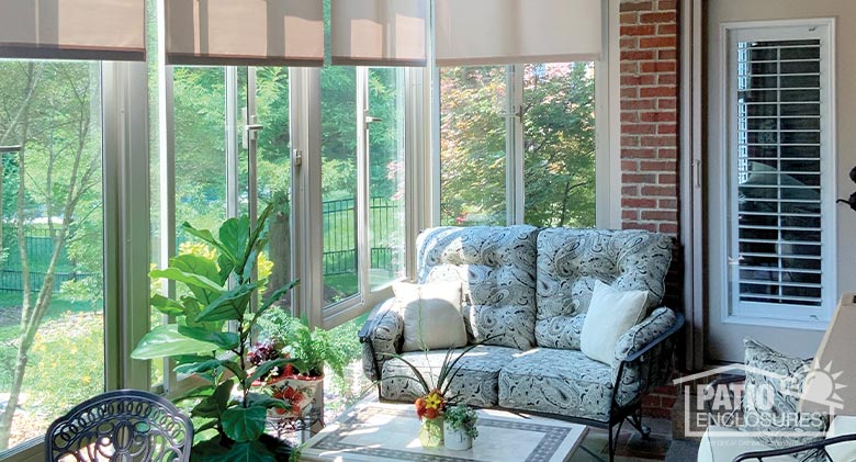 Sun streaming in the windows of a sunroom with open shades, comfortable furniture, a table and plants.