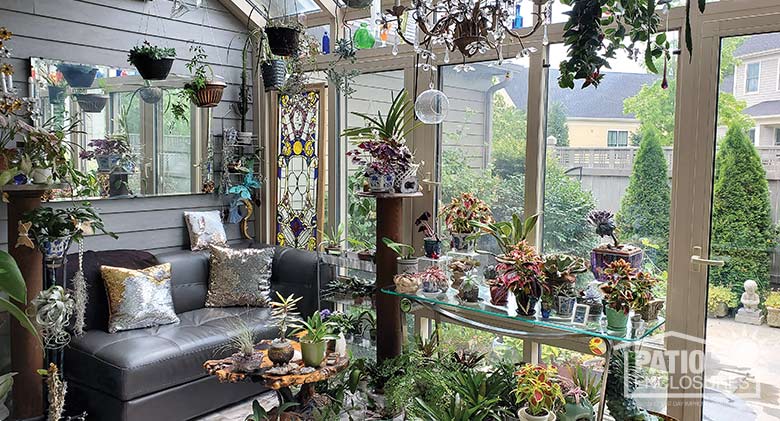 Solarium with leather couch and wide array of plants hanging and on tables, stained glass and colored bottles in the windows.