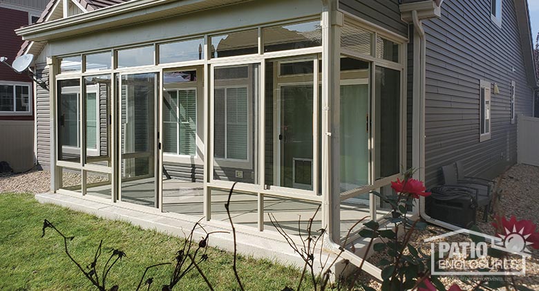 A covered patio enclosed with glass and beige frame attached to a home with gray siding, a rosebush in the foreground.