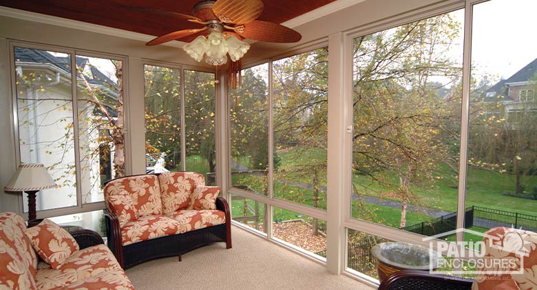 Interior of a second-story sunroom with ceiling fan and wicker furniture with print cushions overlooking a yard and driveway.