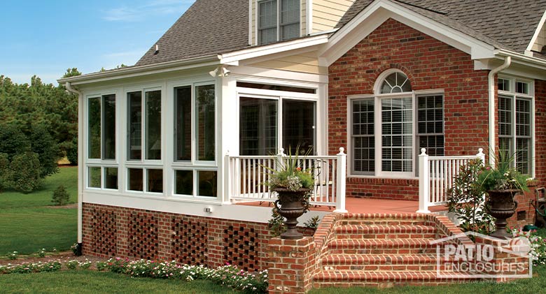 A white glass sunroom encloses part of a raised deck attached to a brick home. Brick stairs with planters descend from deck.