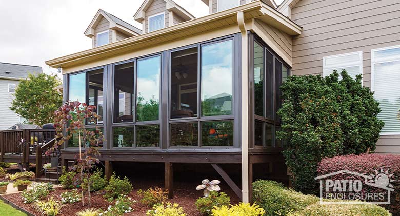A brown sunroom encloses half of a raised deck on a tan home with nice landscaping all around.
