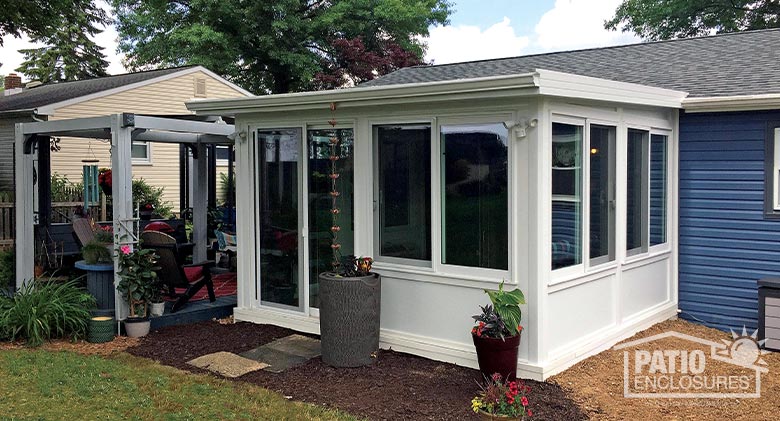 A cute sunroom with solid knee wall next to an open pergola. A rain barrel and rain chain are in front of the sunroom.