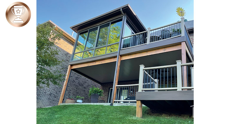 Looking up at a second-story sunroom with brown frame attached to an open deck with stairs leading to lower deck.