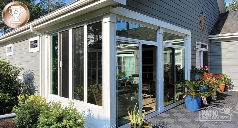 The exterior of a white sunroom enclosing a corner patio with plants and landscaping around it on a gray house.