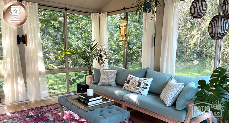 Light blue couch and tufted bench with pillows, hanging lamps, plants and sheer curtains inside a sunroom.
