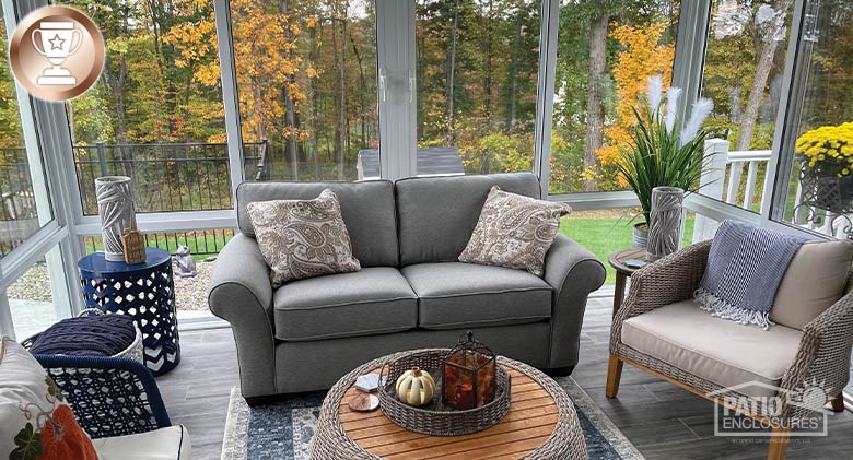 View of colorful trees through sunroom windows. Comfortable furnishings with pillows, a throw and fall décor.