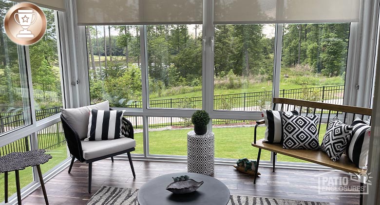 A sunroom with a chair and bench adorned with black and white pillows, a wood floor with rug and partially drawn shades.  