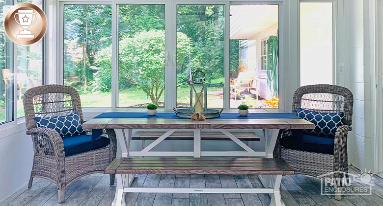 A white sunroom with a solid knee wall, wood floor and a table with two benches and two wicker chairs with blue cushions.
