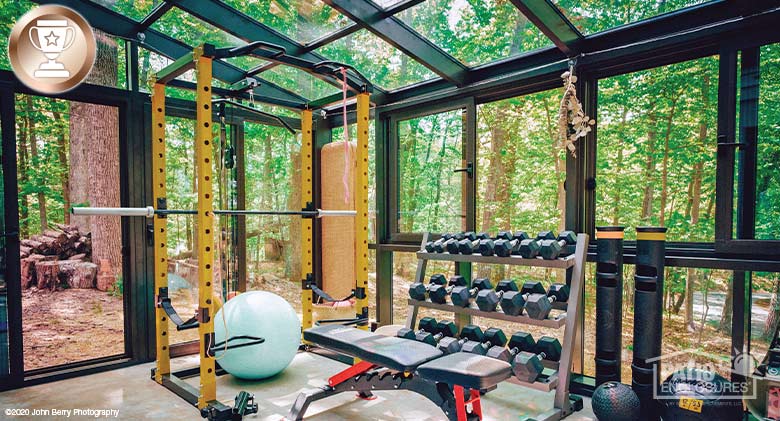 Interior of a bronze solarium with glass roof filled with workout equipment such as weights, bench and stability ball.