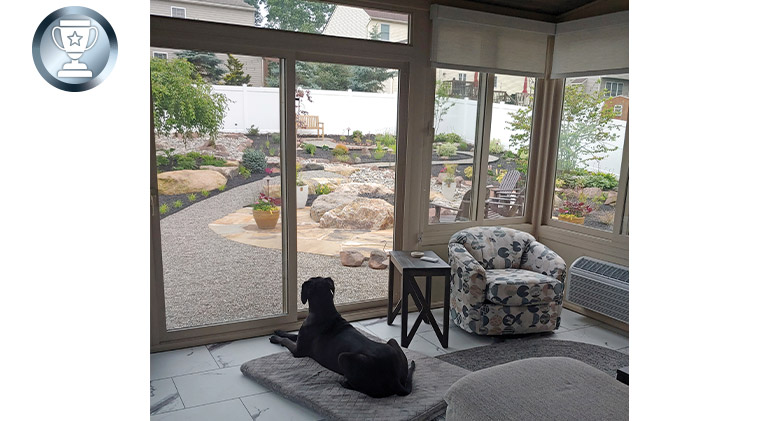A large black dog lays on a cushion looking out of sunroom doors into a nicely landscaped yard with gravel paths and rocks.