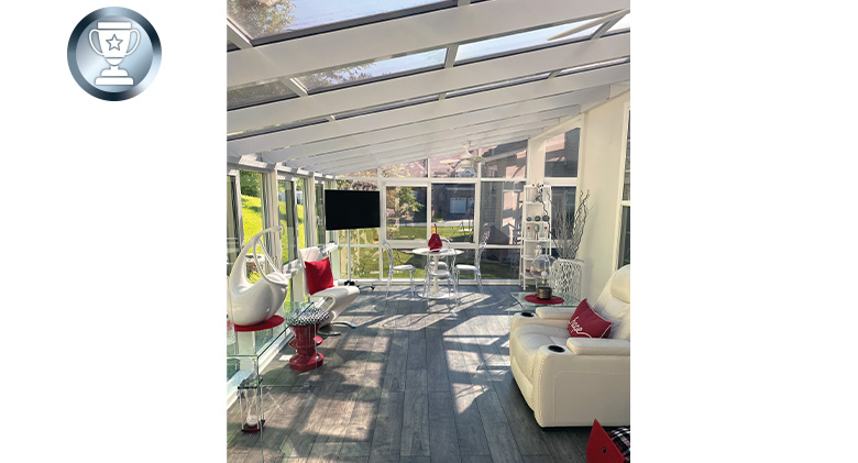 Interior of a white solarium with glass ceiling, wood floor and red and white furnishings. A TV is on a stand in the corner.