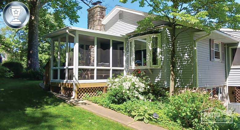 Light gray home with white sunroom, bay window and lush, well-tended garden and trees. A paved walkway leads to the sunroom.
