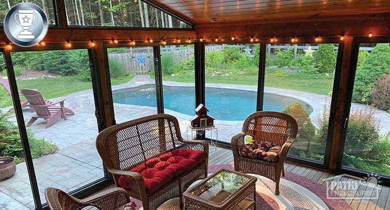 View of a patio and pool from inside a sunroom with brown frame, string lights and wicker furnishings with bright cushions.
