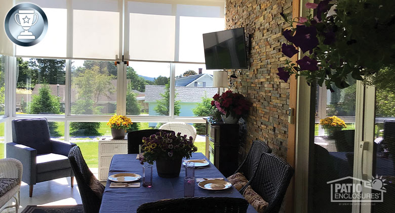 Dining table with places set in the shaded area of a glass sunroom. Two chairs in the sun by windows with half-drawn shades.