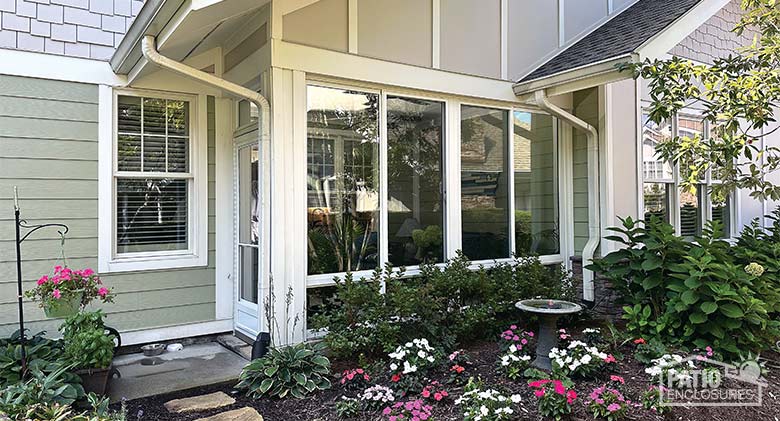  A three-season sunroom porch enclosure with a white frame and glass knee wall, with bright flowers planted in front.
