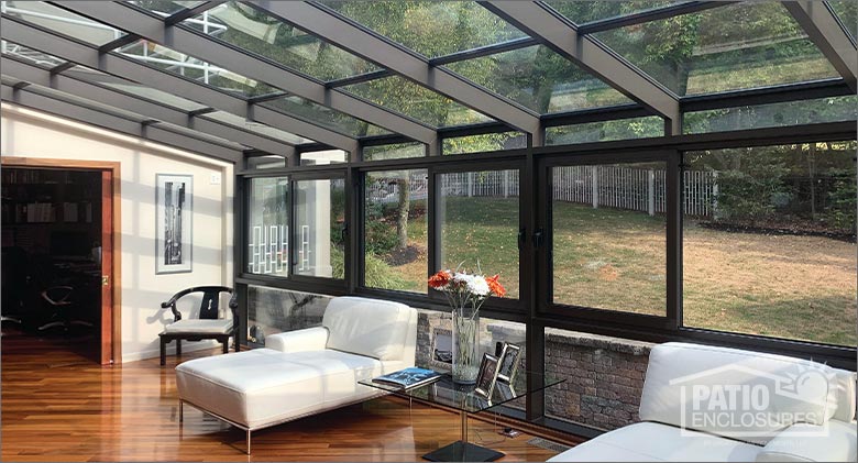 Interior of a brown solarium with glass roof, two white chaise lounges with glass table between them and chair in the corner.