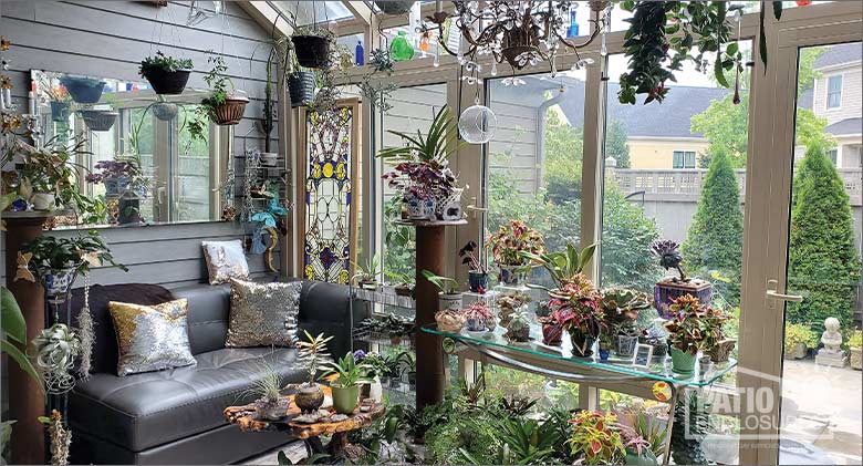 Solarium with many potted plants on a table, on the floor and hanging, a gray couch on left and stained glass in the corner.