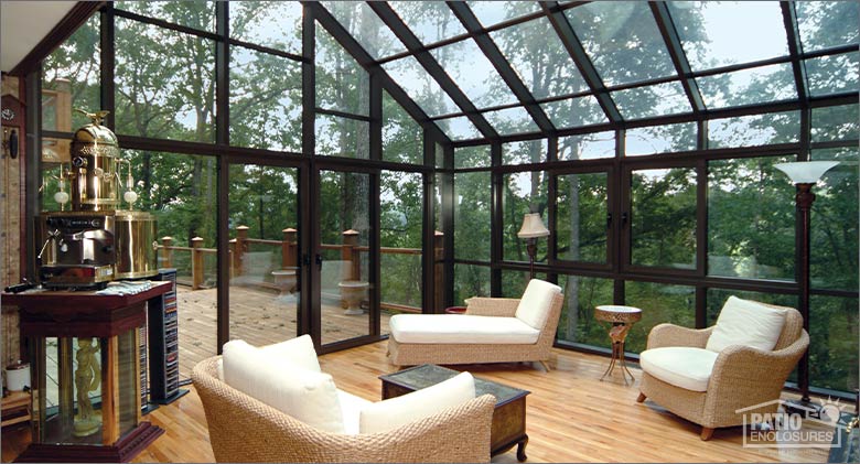 Interior of large solarium with soaring glass ceiling, wicker furniture with white cushions and wood floor. View of woods.