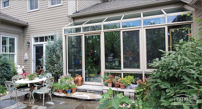 Exterior of a bump-out solarium on enclosed porch of tan house, lush landscaping and a table and chairs on patio outside.