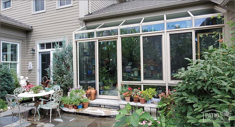  Exterior of a bump-out solarium on enclosed porch of tan house, lush landscaping and a table and chairs on patio outside.