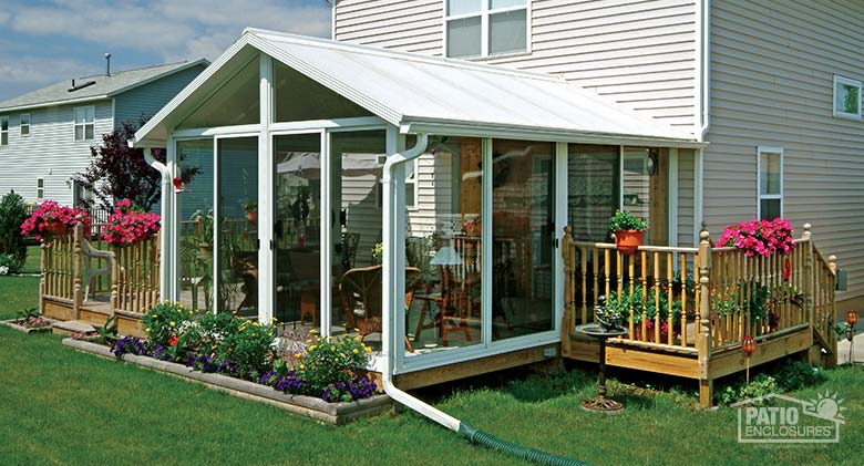 A white sunroom with a gable roof and flowerbed in front of it, a wood deck on each side with pink flowers on the railings.