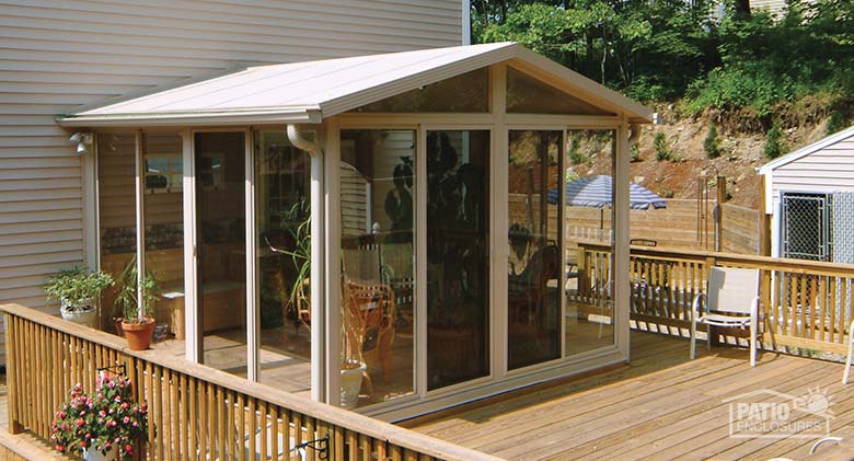 Exterior of a tan sunroom with gable roof built on a wooden deck with railing and pink flowers on the left.