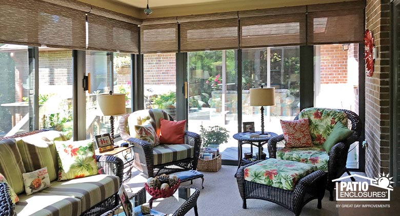 A Florida room with wicker furniture, brightly patterned cushions, table lamps, and brown rolling shades for light control.