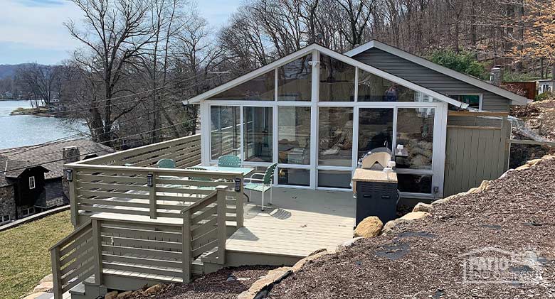 Looking down at a sunroom with a large deck with table, chairs and grill. A body of water in the background.