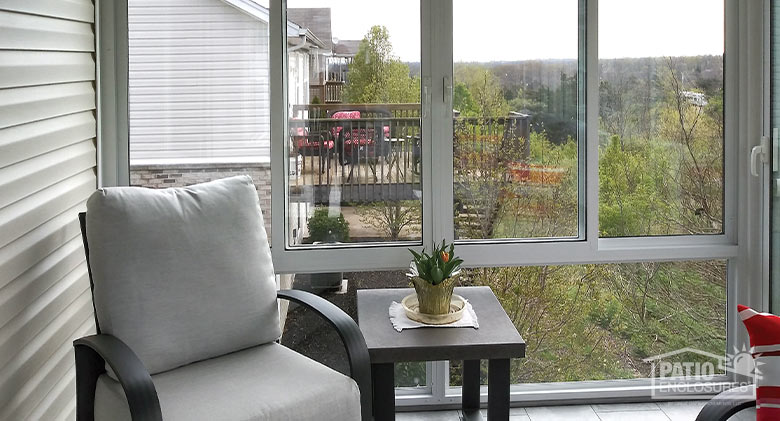 The interior of a sunroom with a comfortable chair and side table and a view of the tops of trees and neighboring homes.