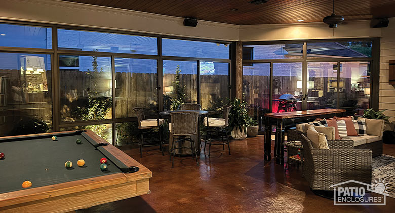 An evening view of a lighted three-season sunroom with billiard table as well as dining and seating areas.